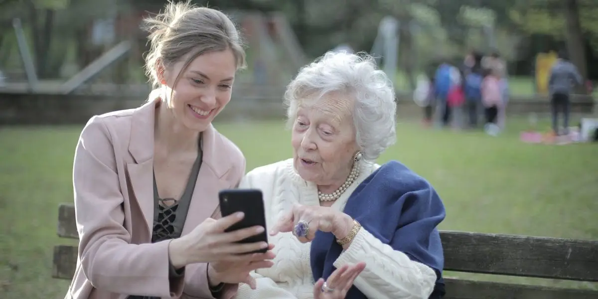 Eine junge Frau zeigt einer älteren Frau etwas auf dem Smartphone, beide sitzen lachend auf einer Bank im Freien