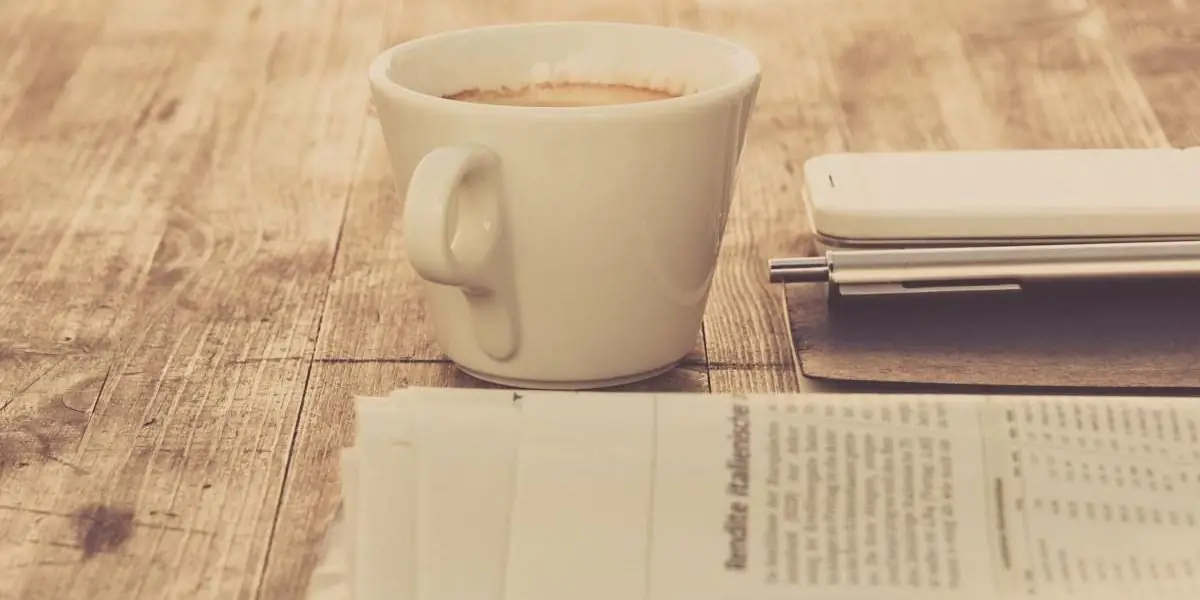 Eine Tasse Kaffee neben einem Stapel Zeitungen und einem Notizbuch auf einem Holztisch